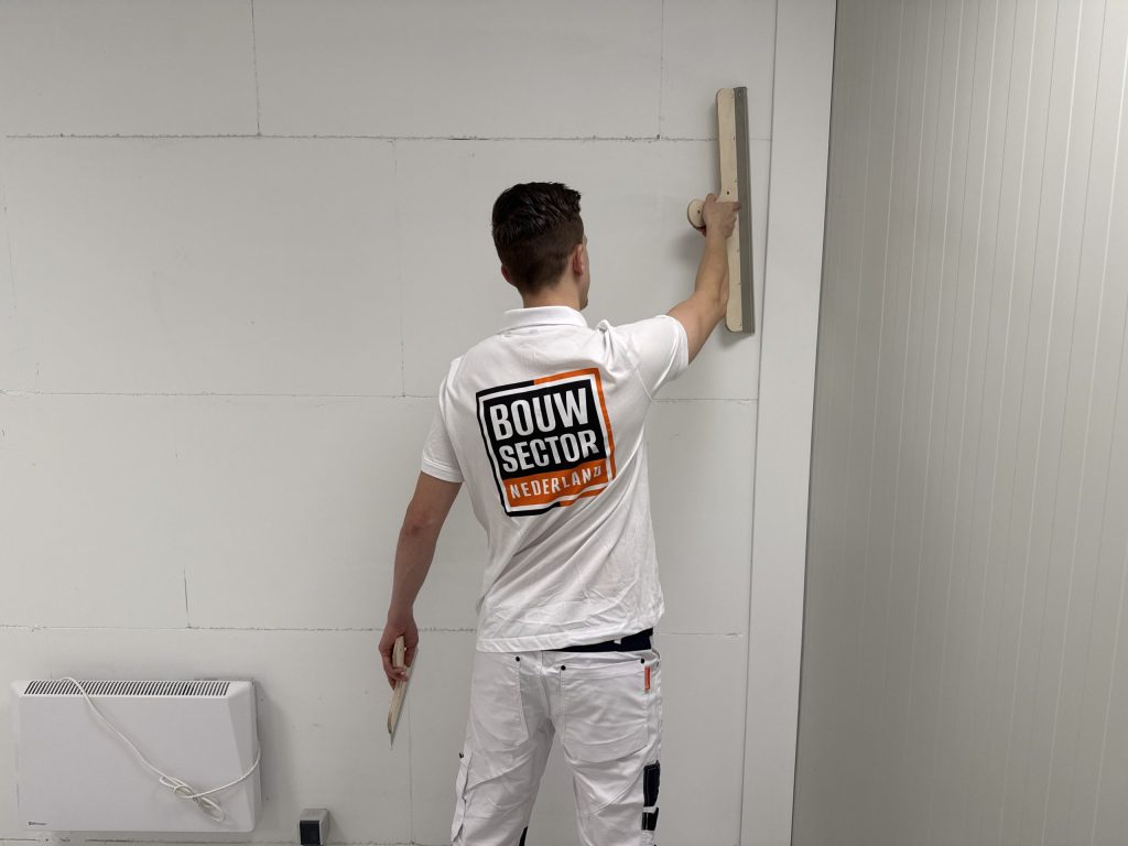 Stukadoor met Bouwsector Nederland-logo op zijn rug strijkt een muur glad met een lange houten spaan