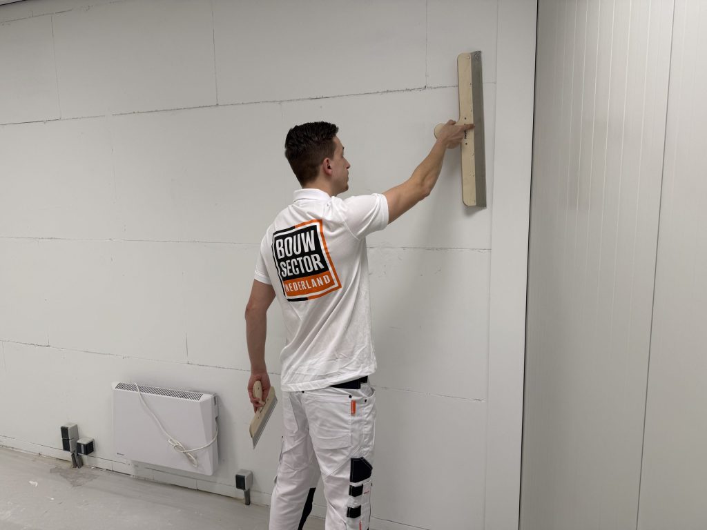 Stukadoor van Bouwsector Nederland zet de spaan tegen de muur van een lege ruimte, klaar voor een sausklare laag