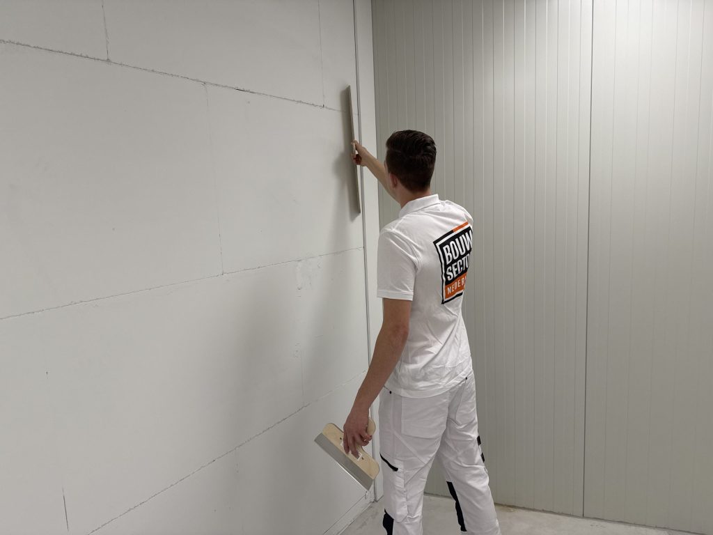Stukadoor van Bouwsector Nederland haalt een lange spaan verticaal langs een muur naast een geribde wand