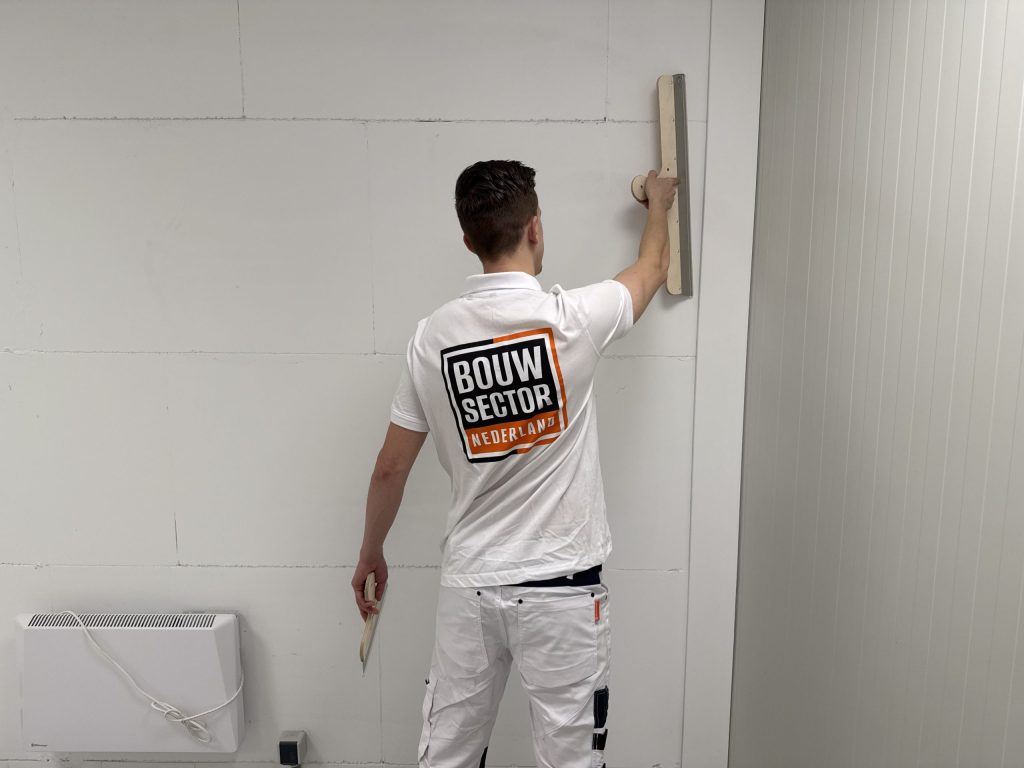 Stukadoor van Bouwsector Nederland in witte werkkleding trekt met de spaan een muur vlak boven een radiator
