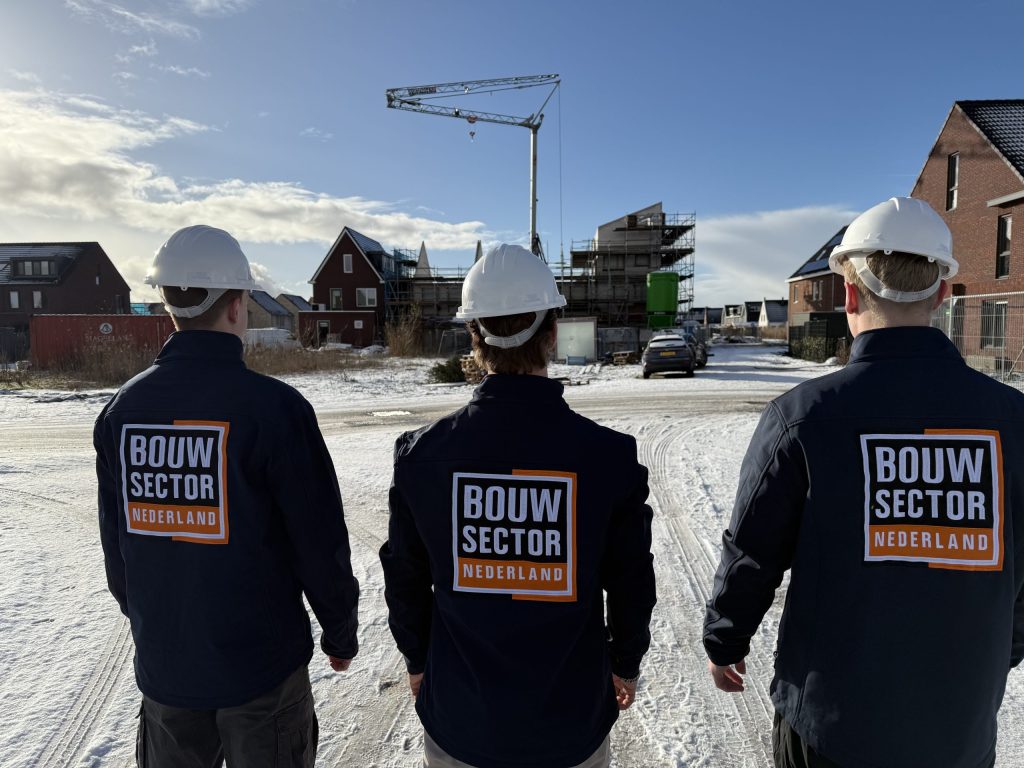 Drie vakmensen van Bouwsector Nederland met witte helm en bedrijfsjas lopen over een besneeuwd bouwterrein