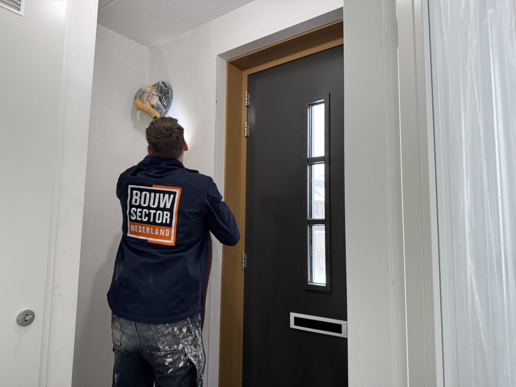 Vakman in donkere Bouwsector-jas schuurt met een wandschuurmachine de muur boven de voordeur in een hal