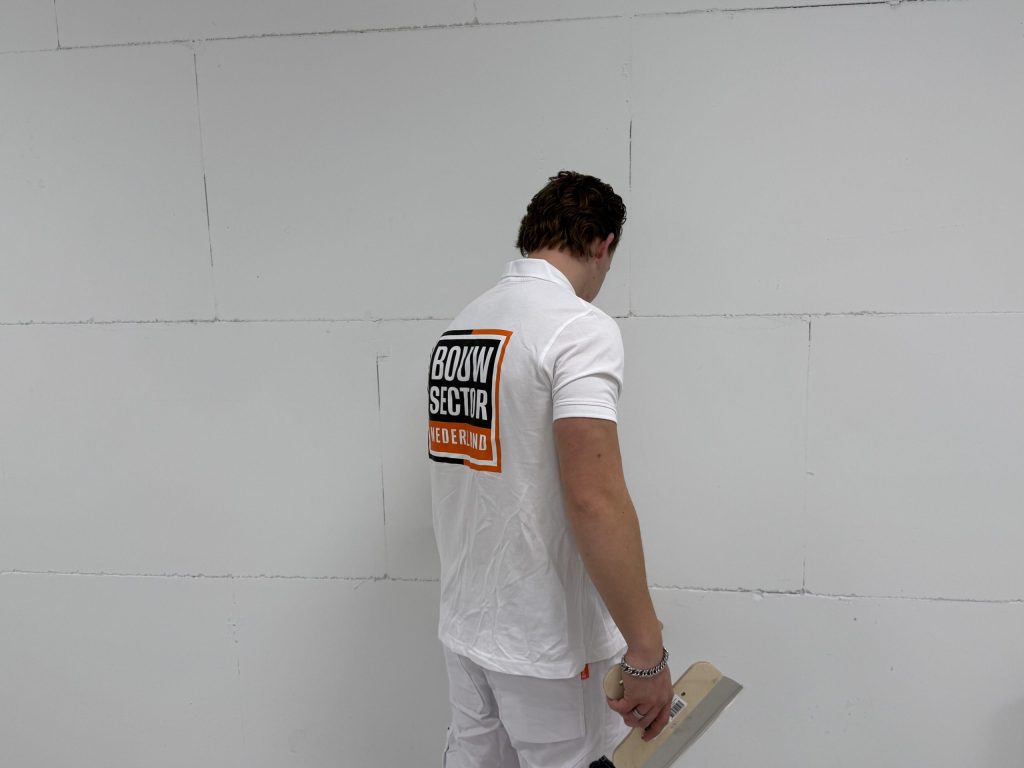 Stukadoor met Bouwsector-logo op zijn rug kijkt naar een kale wand van gipsplaat met zichtbare naden